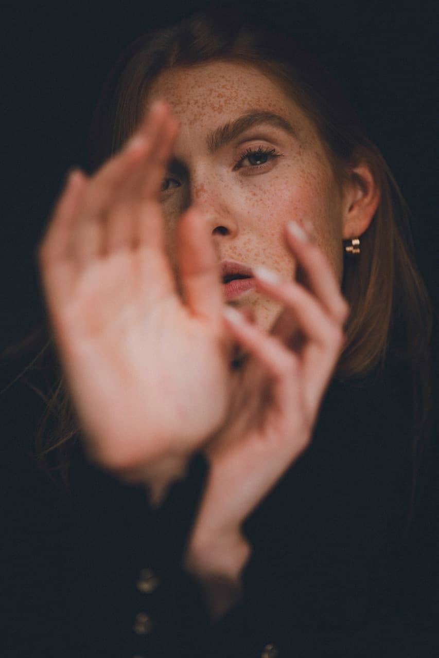 Close-up portrait of Fabia reaching toward the camera with blurred hands in the foreground, her freckled face emerging from a dark background with intense eye contact and dramatic studio lighting.