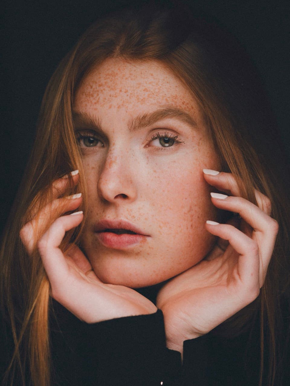 Intimate portrait of Fabia gently holding her face with both hands, looking directly at the viewer. Her freckles are highlighted by soft dramatic lighting against a deep black backdrop.