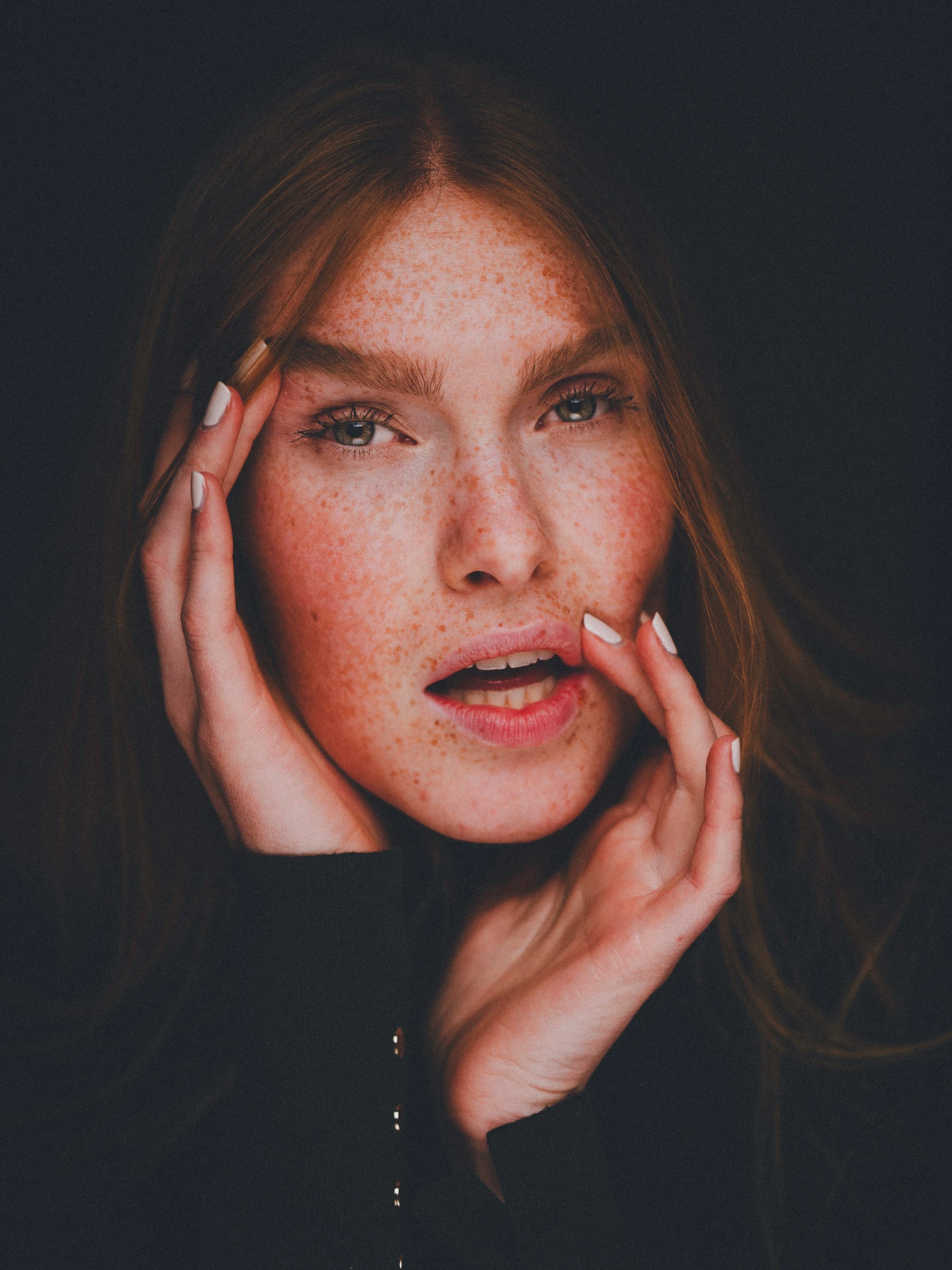 Powerful close-up of Fabia with hands touching her temple and lips, mouth slightly open in an expressive moment. Warm light dramatically illuminates her freckled skin against a dark studio background.